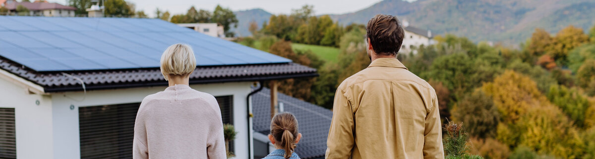 Familie steht vor einem Haus mit Solarpanelen auf dem Dach und genießt die sonnige Umgebung.